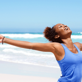 Woman standing on a beach by the water with arms open wide and big smile feeling happy and healthy