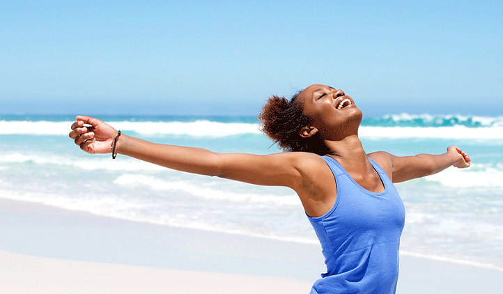 Woman standing on a beach by the water with arms open wide and big smile feeling happy and healthy